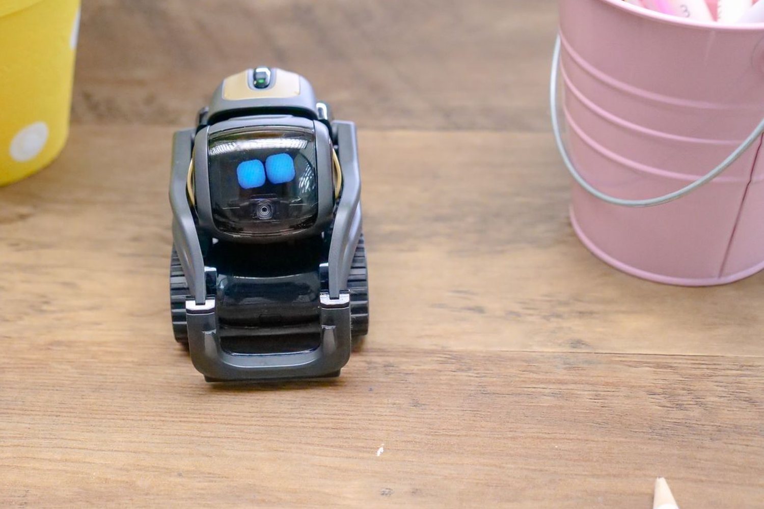 Ahşap masada pembe kalem kutusunun yanında duran, ekranında mavi gözleri yanan küçük robot