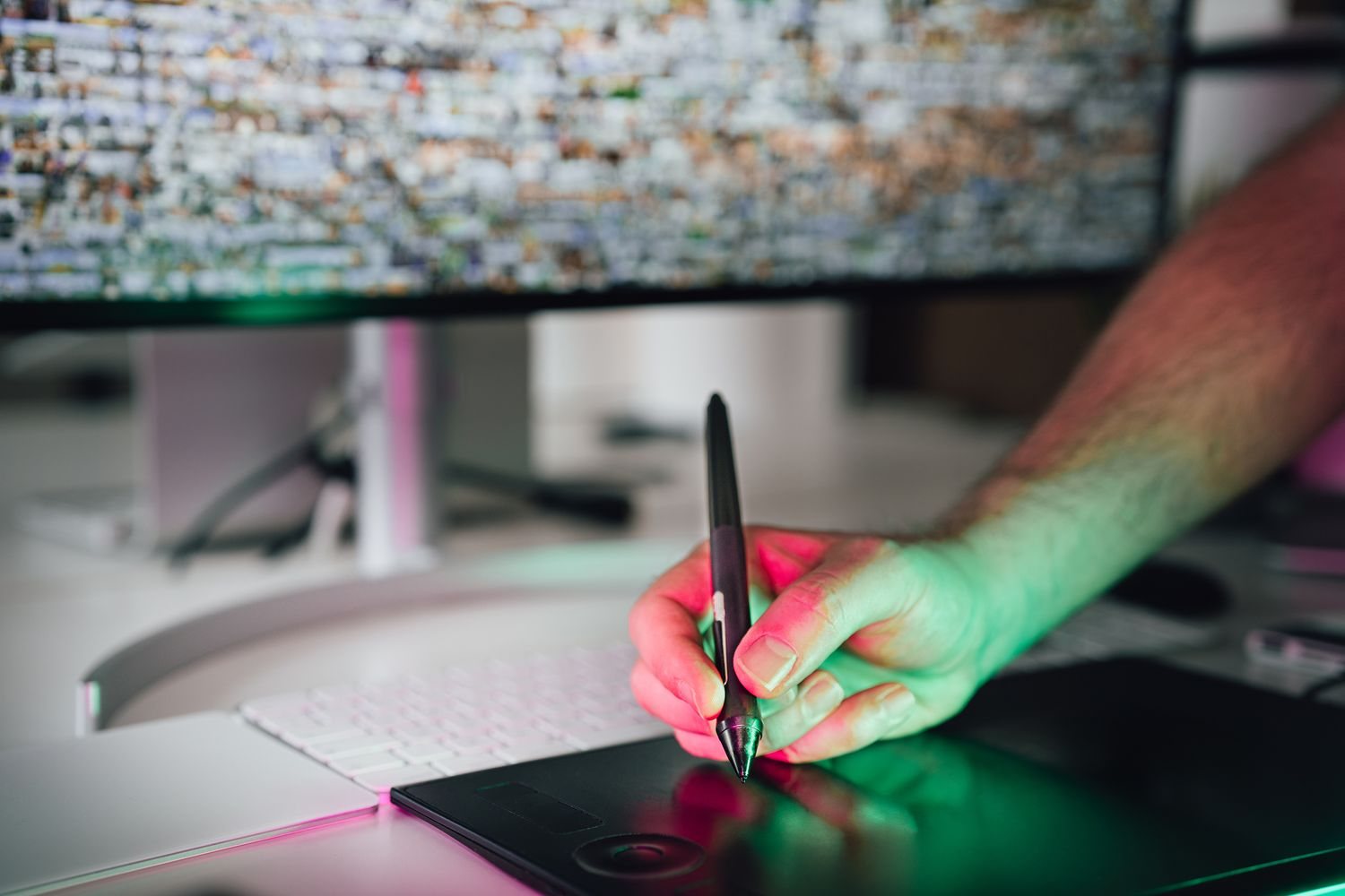 Neon ışık altında grafik tablette kalemle çalışan el; arkadaki ekranda çok sayıda küçük görsel